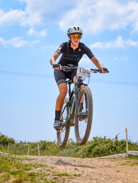 Jenny Bolsom on her mountain bike wearing Saint Piran kit, a helmet and sunglasses. In the photo, her bike is up in the air above the gravel track below as she has just ridden over a bump.
Jenny Bolsom from Truro has been selected as one of Cycling UK's 100 Women in Cycling announced on Thursday 23 November.
Every year the 100 Women in Cycling list celebrates inspirational women who are encouraging others to experience the joy of cycling.
Jenny Bolsom has been nominated as a sporting hero for her work as a Manager of Saint Piran Women Race Team.