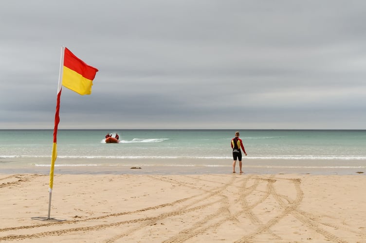 Lifeguards at Mawgan Porth beach, Cornwall. Lifeguards on an arancia inshore rescue boat heading in towards teh beach, lifeguard in wetsuit on shore next to red and yellow flag.